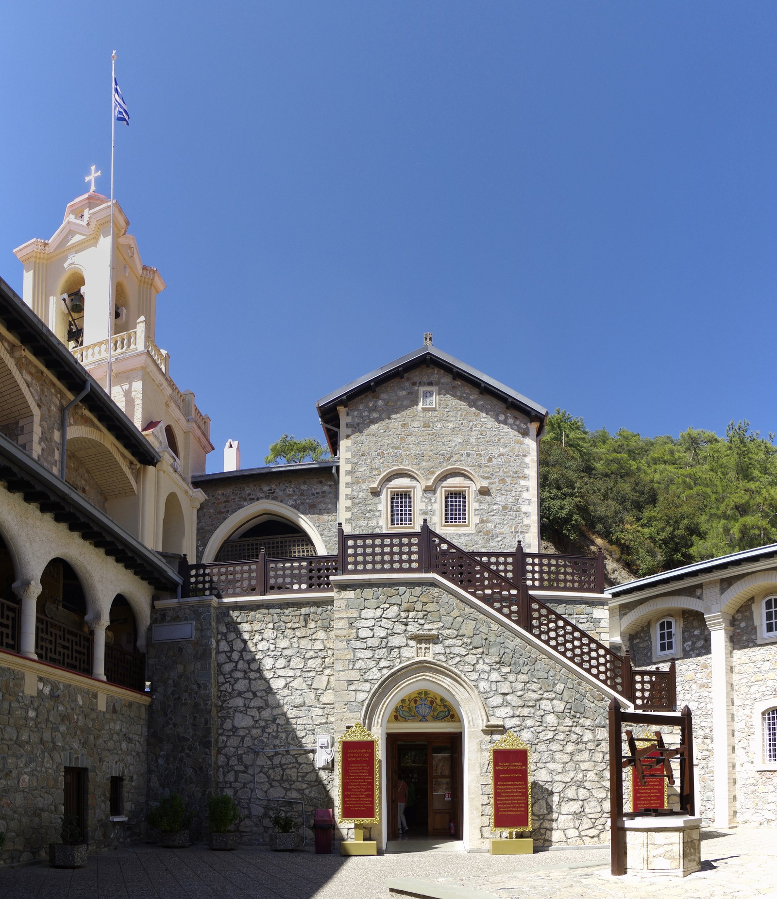 The Holy Monastery of Kykkos in the Troodos mountains — the bell tower with its three crosses and Greek flag rises beside the stone facade, the arched balconies of the corridor, and the carved staircase that climbs to the katholikon.